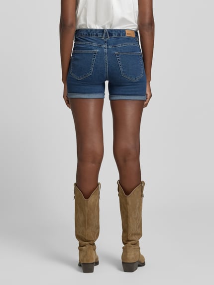 Frauenmodel trägt Regular Fit Jeansshorts aus Baumwoll-Mix Modell 'LUNA' von Vero Moda, Denim-Optik, Label-Patch, 5-Pocket-Design, Beinabschluss mit fixiertem Umschlag, Knopf und verdeckter Reißverschluss, Ganzkörperansicht Rückansicht.
