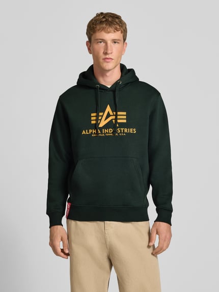 Männermodel trägt Regular Fit Hoodie aus Baumwoll-Mix mit Logo-Print von Alpha Industries, dunkelgrün mit gelbem Print, Ganzkörperansicht von vorne. Rückenlänge 70 cm, Ärmellänge 64 cm, Kapuze mit Tunnelzug, elastische Abschlüsse.