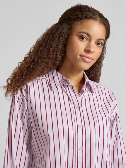 Kobiece modelka nosi Oversized Hemdbluse mit Brusttasche od Gina Tricot z bawełny, krój oversized fit, kołnierzyk typu kent, listwa guzikowa, brustasza, sportowe mankiety, zaokrąglony dół, długość tyłu w rozmiarze S: 71 cm, długość rękawów w rozmiarze S: 54 cm, różowe pionowe paski, tekstura gładka, półzbliżenie górnej części ciała.