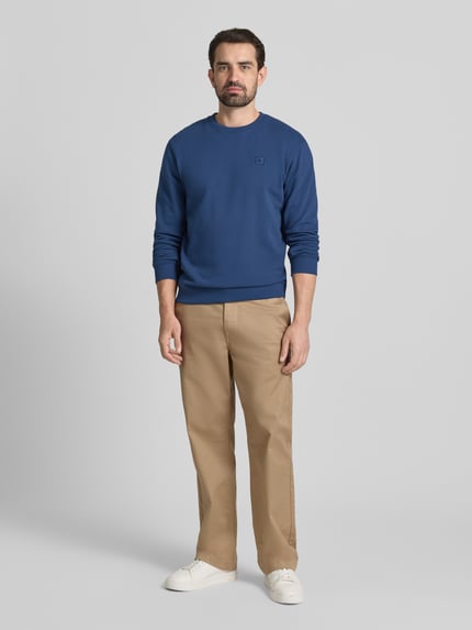 Männermodel trägt Sweatshirt mit Label-Detail von Scotch & Soda in Blau aus reiner Baumwolle, lockerer Schnitt, Ganzkörperansicht von vorne.