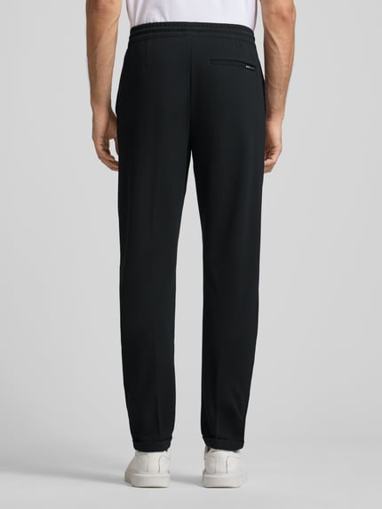 Męski model nosi Tapered Chino mit Viskose-Anteil od Tom Tailor Denim, czarne, gładkie, elastyczny pas, kieszenie, widok całej sylwetki z tyłu.