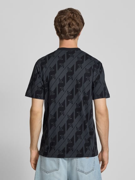 Männermodel trägt T-Shirt aus reiner Baumwolle Modell 'MONOGRAMM AOP' von adidas Originals, dunkle Farben, diagonales Muster, Ganzkörperansicht Rückansicht.
