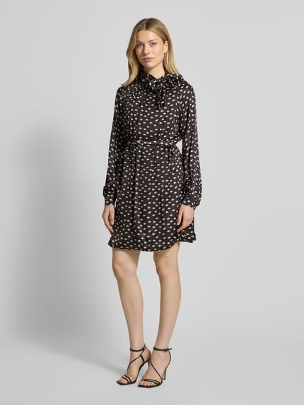 Frauenmodel trägt Cocktailkleid mit Stehkragen von Rinascimento aus reiner Viskose, taillierter Schnitt, Schleifen-Detail, Taillengürtel mit Dornschließe, Rückenlänge bei Größe S 87 cm, Brustweite 61 cm, Ärmellänge 62 cm, schwarz mit beigefarbenem Muster, Ganzkörperansicht von vorne.