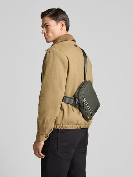 Männermodel trägt Crossbody Bag mit Statement-Print Modell 'EXPLORE' von camel active in Oliv mit Reißverschlussfach, Ganzkörperansicht Rückansicht.