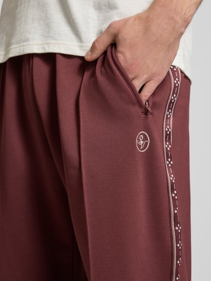 Męski model nosi Trainingshose mit Biese, aufgesetztem Band und Logo-Bestickung von REVIEW, krój regular fit, zaszewka, tasiemka przy szwie bocznym, wyhaftowane logo na nogawce, elastyczny pas, długość wewnętrzna nogawki w rozmiarze M: 43 cm, kolor bordowy, gładka tekstura, detaliczne ujęcie od przodu.