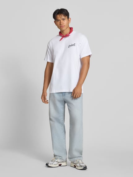 Männermodel trägt Regular Fit T-Shirt aus reiner Baumwolle Modell 'Pup' von Carhartt Work In Progress, weiß, Label-Print, Ganzkörperansicht von vorne. Herren T-Shirt, Rückseitiger Motiv-Print, Rückenlänge bei Größe M: 77 cm.