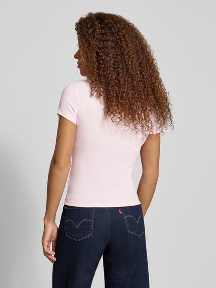Modelka kobieca nosi Slim Fit T-Shirt aus Baumwoll-Mix od Tommy Jeans, jasnoróżowy, krótki rękaw, dekolt V, logo, gładka faktura, widok całej sylwetki z tyłu.