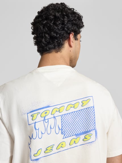 Männermodel trägt Regular Fit T-Shirt aus reiner Baumwolle Modell '90s' von Tommy Jeans, cremefarben mit Label Print, Ganzkörperansicht Rückansicht.