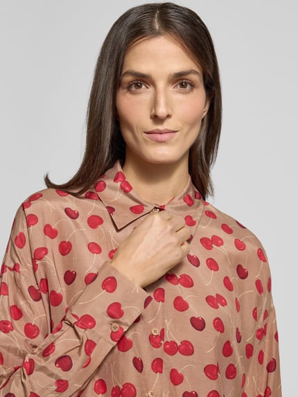 Modelka kobieca nosi Hemdbluse mit Knopfleiste Modell 'CLOELIA' od Drykorn z bawełniano-wiskozowej tkaniny, wzór w czerwone wiśnie, luźny krój, koszulowy kołnierzyk, zapięcie na guziki, kolor naturalny z czerwonym. Zbliżenie na górną część ciała.