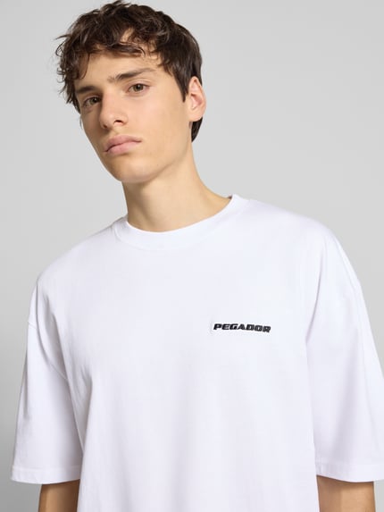 Mannelijk model draagt Oversized T-Shirt met Label-Stitching van Pegador in wit, pure katoen, geribde ronde hals, borstlogo, close-up bovenlichaam.