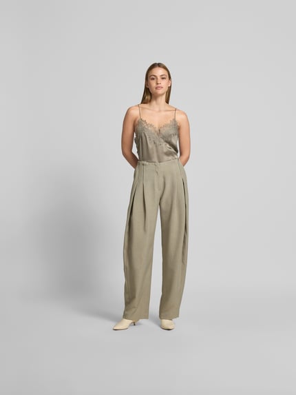 Frauenmodel trägt Wide Fit Stoffhose mit Bundfalten von Iro in Beige, mit Eingrifftaschen, Gesäßtaschen, Haken- und Reißverschluss; Ganzkörperansicht von vorne.