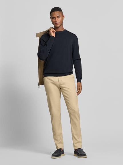 Męski model nosi Slim Fit Chino aus Baumwoll-Mix von Tommy Hilfiger, jednokolorowy, krój slim fit, piqué, mieszanka bawełny, z wyhaftowanym logo, wpuszczane kieszenie, guziki, zamek, widok całej sylwetki z przodu.