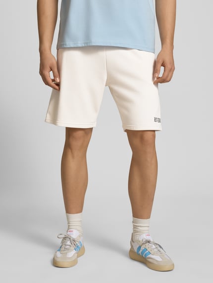 Männermodel trägt Regular Fit Shorts mit Label-Stitching von REVIEW in Creme aus Baumwoll-Mix, elastischer Bund, Nahaufnahme Beine, Ganzkörperansicht von vorne. Bundweite 44 cm, Innenbeinlänge 22 cm, Regular-Fit Herren Shorts.