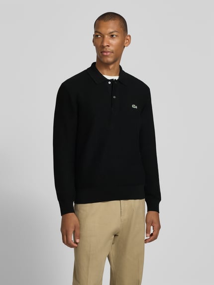Męski model nosi czarną koszulkę polo Regular Fit Poloshirt aus reiner Baumwolle od Lacoste, beżowe spodnie, całkowity widok z przodu.