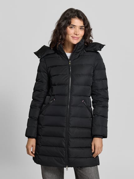 Frauenmodel trägt Daunenmantel mit Kapuze von Tommy Hilfiger in Schwarz mit Logo-Detail, Zweiwege-Reißverschluss und Reißverschlusstaschen, Longline-Schnitt, Webware mit Steppeinsatz, Ganzkörperansicht von vorne.