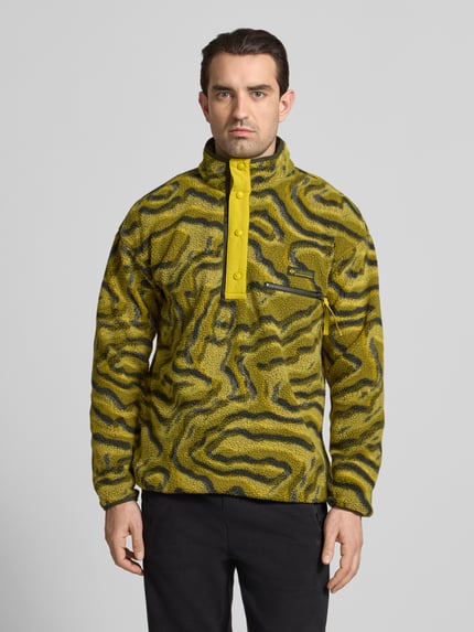 Männermodel trägt Regular Fit Pullover mit Logo-Patch von Columbia in gelber Fließ-Optik mit Brusttasche, Druckknopfleiste, Ganzkörperansicht von vorne.