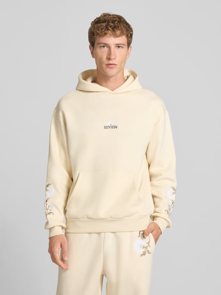 Männermodel trägt Relaxed Fit Hoodie mit Känguru-Tasche von REVIEW in Creme mit floraler Stickerei, Baumwoll-Mix, Nahaufnahme Oberkörper.