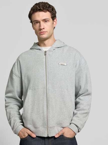 Männermodel trägt graue Regular Fit Sweatjacke mit Känguru-Tasche Modell 'L’ATELIER LINE' von RETERNITY, Ganzkörperansicht von vorne. Baumwoll-Elasthan-Mix, Kapuze, Reißverschluss, Label-Detail sichtbar.