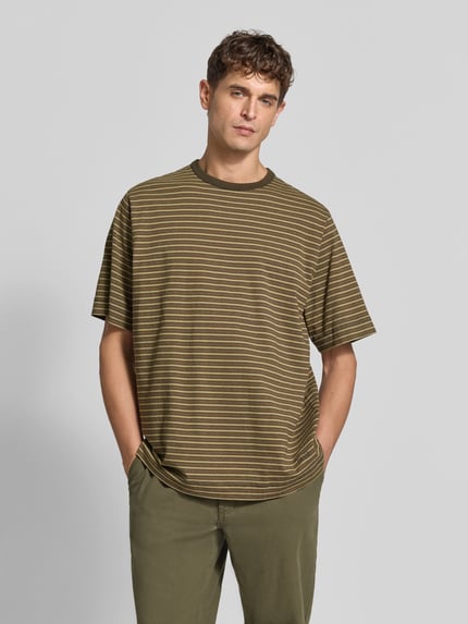 Männermodel trägt Relaxed Fit T-Shirt mit Label-Stitching von Forét, Streifenmuster in Oliv und Weiß, Rundhalsausschnitt, Halbarm, reine Baumwolle, Rückenlänge 73 cm bei Größe M, Ganzkörperansicht von vorne.