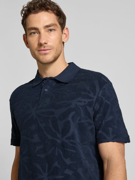Männermodel trägt Regular Fit Poloshirt aus Baumwoll-Mix Modell 'PE_FLOWER' von BOSS Orange in Dunkelblau mit Allover-Muster, Nahaufnahme Oberkörper.
