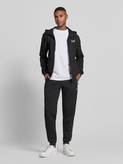 Männermodel trägt schwarze Sweatpants mit Viskose-Anteil Modell 'TRAIN PREMIUM SHIELD' von EA7 Emporio Armani, Gerade Passform, Reißverschlusstaschen, Gerippte Abschlüsse, Elastischer Bund, Elastischer-Material-Mix, Ganzkörperansicht von vorne.