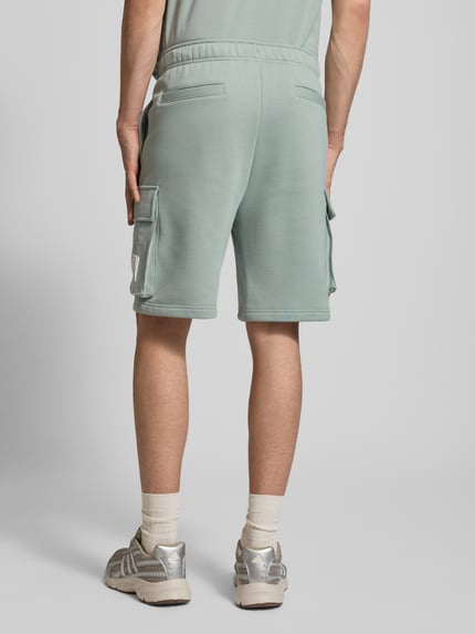 Männermodel trägt Sweatshorts mit Cargotaschen und Logo von CHAMPION in Mintgrün, Baumwoll-Mix, Relaxed Fit, Ganzkörperansicht Rückansicht.