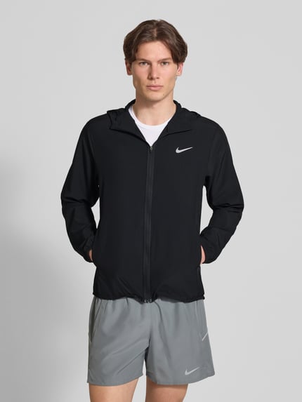 Männermodel trägt zwarte trainingsjack met logoprint en opstaande kraag, ritssluiting, zijsteekzakken, elastische mouwboord, recht model, ruglengte bij maat M 69 cm, mouwlengte 65 cm, 'Trainingsjacke mit Logo-Print und Stehkragen' van NIKE TRAINING, Ganzkörperansicht van voren.