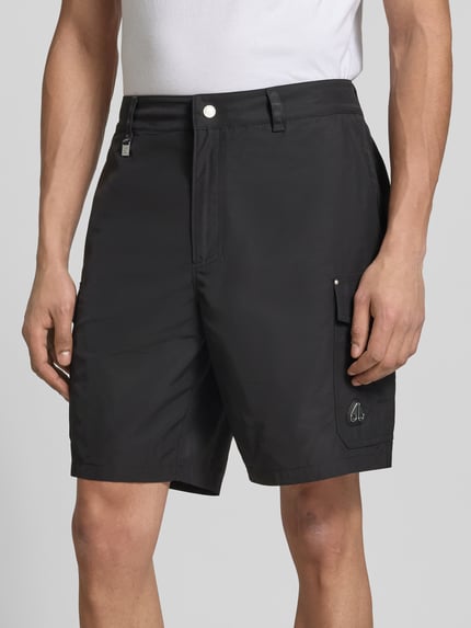 Mężczyzna nosi Regular Fit Shorts mit Cargo-Taschen od Moose Knuckles, czarne, krój regular fit, kieszenie cargo, wpuszczane i z tyłu, detal z logo, zapięcie na guzik i zamek błyskawiczny, długość wewnętrzna nogawki w rozmiarze M: 21 cm, szerokość w rozmiarze M: 45 cm, detal: krótki przód, całkowite ujęcie od przodu.