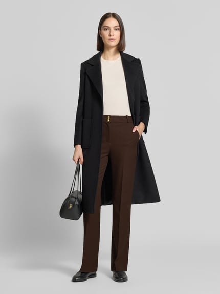 Modelka nosi Slim Fit Schmale Hose mit Viskose-Anteil od GANT w jednolitym brązowym kolorze, Kanty, wpuszczana kieszeń, splot twill, dodatek wiskozy; krój slim fit, średni stan, zapięcie na guzik i kryty zamek błyskawiczny, widoczne detale kieszeni, wykończenie materiału: twill, widoczne jednolite faktury, Ganzkörperansicht von vorne.