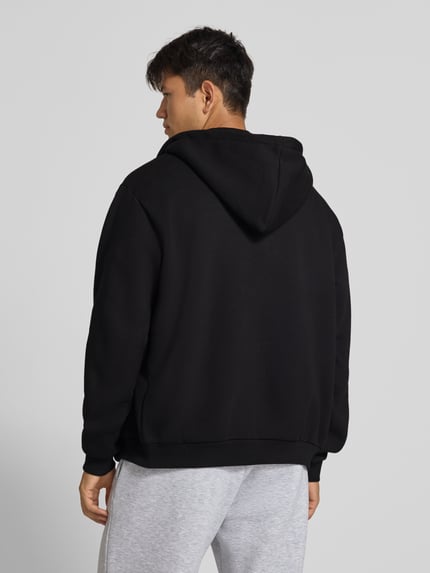 Männermodel trägt Sweatjacke mit Label-Stitching und Känguru-Tasche von Karl Kani im Baumwoll-Mix, Schwarz, Regular Fit, Ganzkörperansicht Rückansicht.