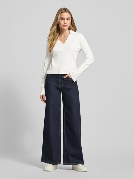Frauenmodel trägt Slim Fit Langarmshirt aus reiner Baumwolle von Tommy Hilfiger in Unifarben mit V-Ausschnitt und Logo-Detail, Ganzkörperansicht von vorne.