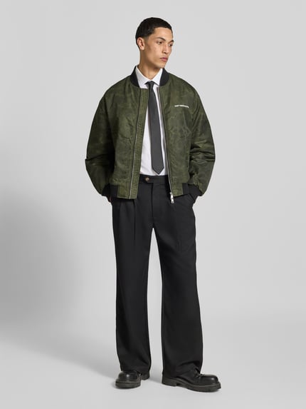 Męskamodel nosi bomberkę 'Bomberjacke mit Label-Print' od Don't Waste Culture w kolorze khaki z nadrukiem z logo, krój regular fit, zamek błyskawiczny dwustronny, elastyczne wykończenia, stójka, długość tyłu w rozmiarze M: 67 cm, szerokość ramion w rozmiarze M: 53 cm, długość rękawów w rozmiarze M: 64 cm, widok całościowy z przodu.