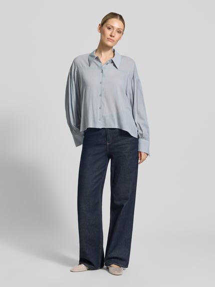 Frauenmodel trägt Relaxed Fit Bluse Modell 'SELENI' von Drykorn aus Baumwoll-Viskose-Mix, hellblau mit Hemdblusenkragen und Knopfleiste, Ganzkörperansicht von vorne. Rückenlänge bei Größe 36: 59 cm, Ärmellänge 63 cm.