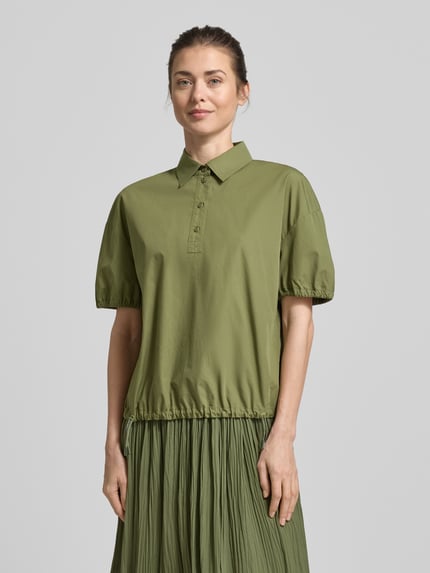 Frauenmodel trägt Regular Fit Bluse mit Polokragen von Marc Cain in Olivgrün, reine Baumwolle, elastische Ärmelabschlüsse, Bund mit Tunnelzug, kurzärmeliges Design, Ganzkörperansicht von vorne.