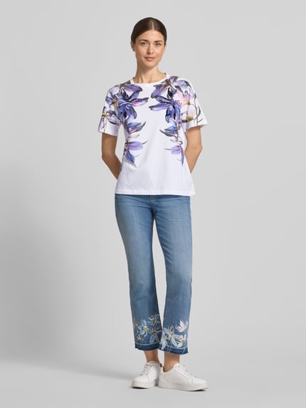 Frauenmodel trägt Damen T-Shirt im Allover-Look mit Pailletten von Marc Cain aus Reine Baumwolle, Regular-Fit, weiß mit violetten Blumen, Rundhalsausschnitt, Rückenlänge 59 cm bei Größe 36, Ganzkörperansicht von vorne.