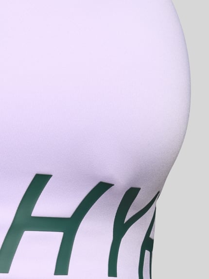 Frauenmodel trägt Sport-BH mit Label-Print von PUMA Performance in hellem Fliederton mit dunkelgrüner Aufschrift, Nahaufnahme Oberkörper.