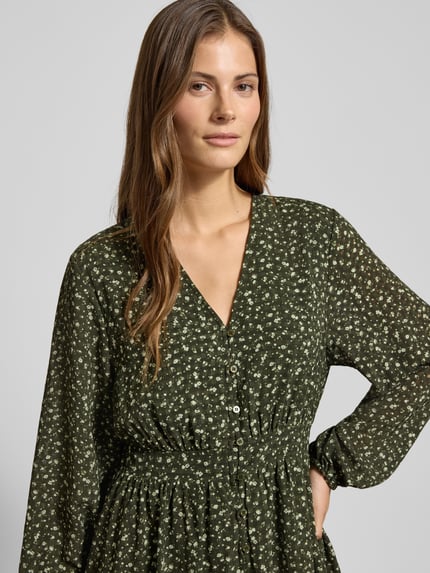 Frauenmodel trägt Minikleid im Allover-Look mit Knopfleiste Modell 'Kaylen Oceanna' von MSCH Copenhagen, Nahaufnahme Oberkörper. A-Linie, V-Ausschnitt, dunkelgrüner Stoff mit feinem Blumenmuster, strukturierte Textur, lange Ärmel.
