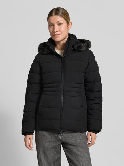 Frauenmodel trägt schwarze Jacke mit abnehmbarer Kapuze Modell 'ECLIPSE' von NAUTGÅRD, gesteppte Struktur, Ganzkörperansicht von vorne. Rückenlänge, Schulterbreite, Brustweite und Ärmellänge bei Gr. S im Kontext: 65 cm, 41 cm, 51 cm, 64 cm. Zweiwege-Reißverschluss, Label-Patch, seitliche Eingrifftaschen mit Druckknopf, gestrickte Ärmelabschlüsse.