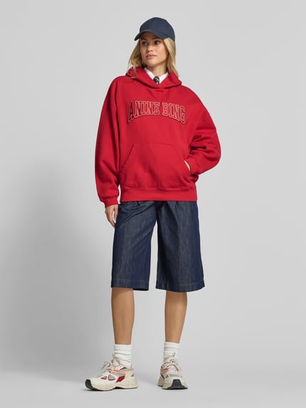 Vrouwenmodel draagt Oversized Hoodie aus reiner Baumwolle van Anine Bing in rood met labelprint, kangoeroezak, ribboorden, denim bermuda, sneakers, complete voorkant gezichtsopname, dames hoodie, lengte 65 cm bij maat S.