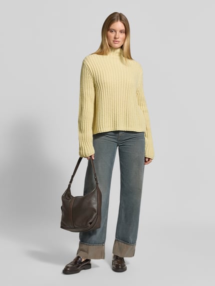 Frauenmodel trägt Strickpullover mit Stehkragen Modell 'ARWIE' von DRYKORN in Gelb, grobe Rippstruktur, kombiniert mit Jeans und dunkler Tasche, Material-Mix mit Alpaka-Anteil, Regular Fit, Ganzkörperansicht von vorne.