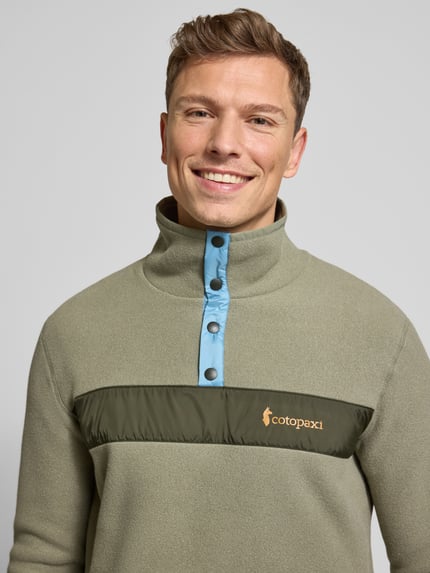 Męski model nosi Sweatshirt mit Label-Print od Cotopaxi w kolorze khaki z niebieską listwą i nadrukiem na piersi, zbliżenie na górę ciała.