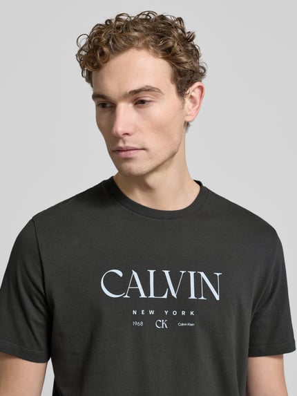 Männermodel trägt T-Shirt mit Label-Print und Rundhalsausschnitt von CK Calvin Klein in Schwarz, reine Baumwolle, Regular-Fit, Nahaufnahme Oberkörper.