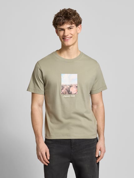 Männermodel trägt T-Shirt mit Rundhalsausschnitt Modell 'JORNORREBRO' von Jack & Jones aus reiner Baumwolle in hellem Khaki mit Frontprint, gerader Schnitt, Ganzkörperansicht von vorne.