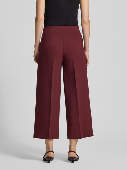Frauenmodel trägt Stoffhose mit Bügelfalte und Eingrifftaschen von MORE & MORE in Bordeaux, glatter Stoff, Ganzkörperansicht Rückansicht. Wide Leg Fit, Material-Mix mit Viskose-Anteil, seitlicher Nahtreißverschluss, französische Eingrifftaschen.