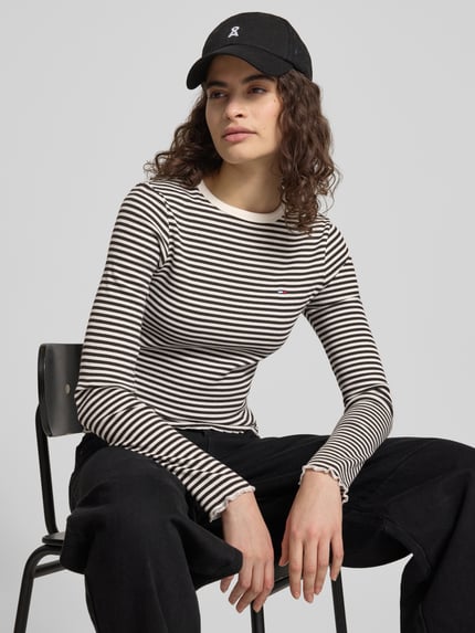 Vrouwenmodel draagt 'Slim Fit Langarmshirt aus Baumwoll-Mix' van Tommy Jeans, gestreept, logo-detail, ronde hals, lange mouwen, frill afwerking, fijnrib, katoenmix. Halfbody vooraanzicht.