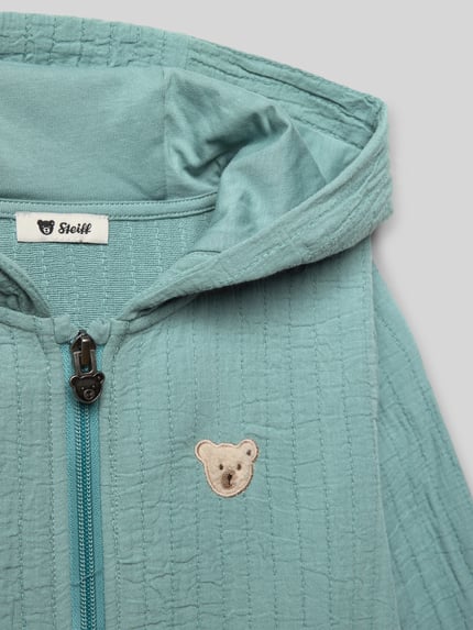 Mädchen trägt Sweatjacke mit Label-Stitching von Steiff, reine Baumwolle, Regular-Fit, Kapuze, Reißverschluss, Känguru-Tasche, Nahaufnahme Oberkörper.