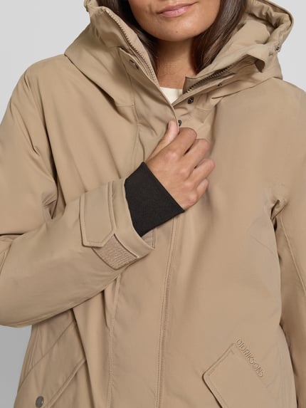 Kobieta nosi Regular Fit Parka mit Kunstfellbesatz Modell 'ERNA' od Didriksons, beżowy kolor, zapinana na suwak, kieszenie z klapką, detal logo, Thermore wypełnienie, wodoodporność 10.000mm, kaptur, szerokość w rozmiarze 36: 51 cm, długość tyłu: 93 cm, szerokość ramion: 42 cm, długość rękawa: 66 cm, zbliżenie na górną część ciała.