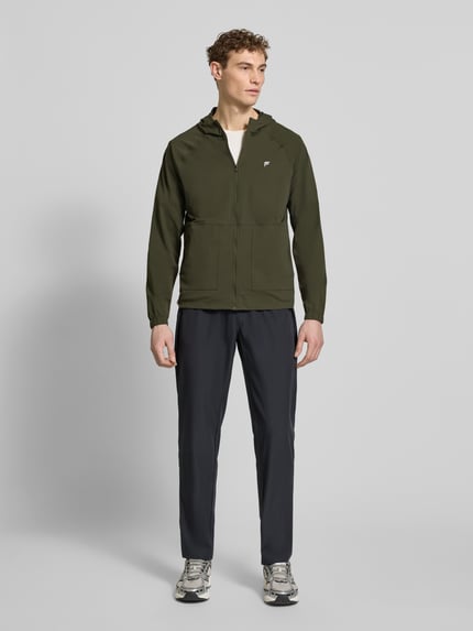 Męski model nosi Regular Fit kurtkę z kapturem model 'The One' od Fabletics, elastyczna mieszanka materiałów, nakładane kieszenie, nadruk z logo, zamek błyskawiczny, kolor khaki i czarny, krój regular fit, całe ciało z przodu