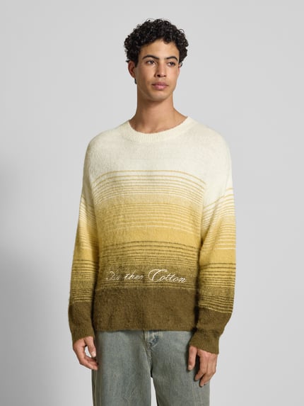 Männermodel trägt Oversized Strickpullover mit gerippten Abschlüssen von ANOTHER COTTON LAB im Farbverlauf Gelb-Oliv, Nahaufnahme Oberkörper, Material-Mix mit Baumwoll-Anteil, Rundhalsausschnitt und Label-Stitching.