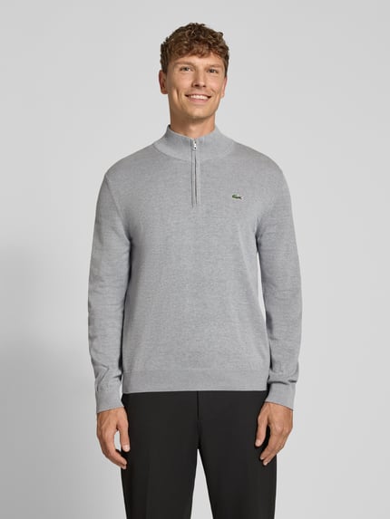 Männermodel trägt Pullover mit halblangem Reißverschluss von LACOSTE in Hellgrau, Feinstrick, Logo-Detail, Ganzkörperansicht von vorne.